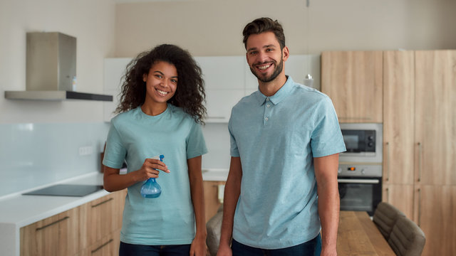 Making Your Home Sparkling Clean. Happy Male And Female Professional Cleaners In Uniform Holding Cleaning Supplies, Looking At Camera And Smiling While Standing In The Kitchen. Cleaning Service