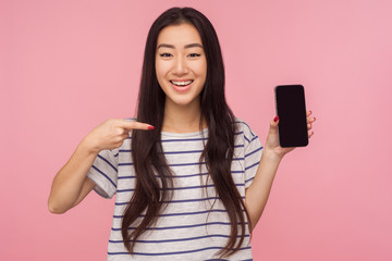 Advertisement of mobile device or application, Portrait of brunette happy girl pointing at cell phone and smiling at camera, satisfied with cellular. indoor studio shot isolated on pink background