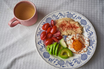 Fototapeta na wymiar Low carb or ketogenic dish consist from fresh cherry tomatoes, avocado, fried egg and chickpea hummus served on rustic plate with cup of the organic green tea. Balanced dish full of proteins and fibre