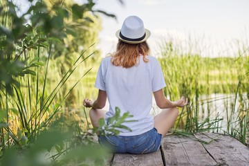 Young woman sitting in lotus position on bridge near water, back view