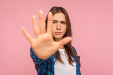 I refuse! Portrait of serious determined girl in checkered shirt showing stop with raised palm, doing negative gesture, warning sign to deny proposal. indoor studio shot isolated on pink background