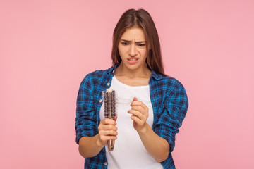 Portrait of stressed brunette woman in casual style clothes looking at unhealthy hair on brush, checking dandruff, upset by hair loss problem, alopecia. indoor studio shot isolated on pink background