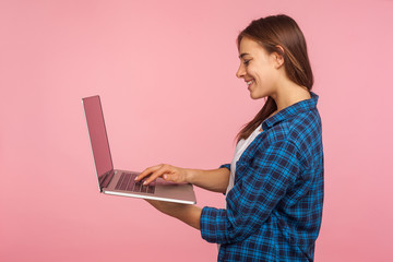 Side view of smiling girl in casual checkered shirt doing freelance task on laptop, using computer for education or online job, browsing internet. indoor studio shot isolated on pink background