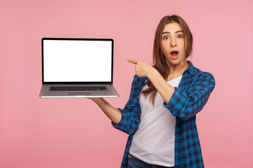 Portrait of amazed charming girl in checkered shirt showing blank laptop screen with shocked...
