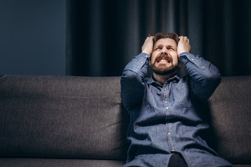 Front view of lonely bearded man in jeans shirt and black trousers sitting without lights at home and suffering from depression. Concept of sorrow and mental pain