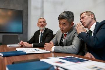 Discussing contract conditions. Caucasian mature businessman whispering something to asian colleague in his ear while having a meeting in the modern office. Business people working together