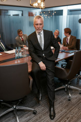 Business expert. Serious mature businessman in formal wear keeping one hand in pocket and looking at camera while leaning at the desk in office