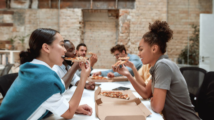 Time for a lunch. Happy multicultural team eating pizza and communicating with each other while sitting together in the modern office
