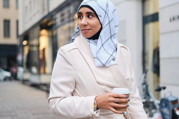 Arabic young casual woman in hijab drinking coffee during walk through street