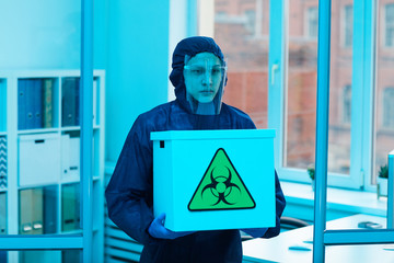 Waist up portrait of male scientist wearing biohazard gear holding box with danger sign while...