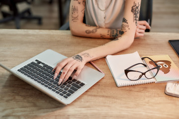 Cropped photo of tattooed woman typing something on the laptop while working in the office