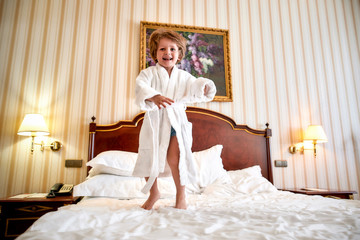 Keeping you moving. Cheerful boy in white bathrobe is having fun, jumping on the white bed in a hotel room. The kid laughs and jumps - obrazy, fototapety, plakaty