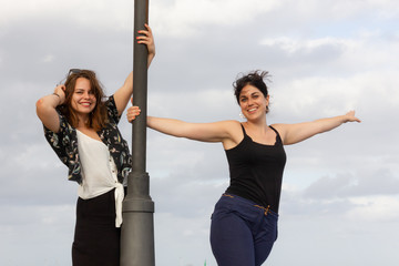 Happy lady friends holding to street lamp on cloudy day. Young women standing with smile. Female...
