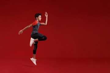 Fototapeta na wymiar Cardio training. A teenage boy is engaged in sport, he is looking aside while jumping. Isolated on red background. Fitness, training, active lifestyle concept. Horizontal shot