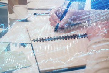 Hand taking notes in notepad. Forex chart holograms in front. Concept of research. Double exposure