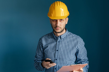 Portrait of young engineer or building contractor wearing yellow protective helmet and shirt standing in front of blue wall background holding smart phone and clipboard looking to camera