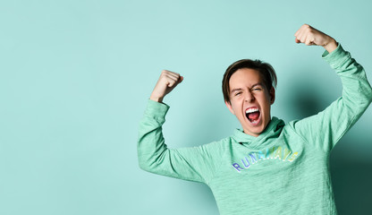 Young teen boy in blue hoodie standing, shouting and feeling powerful over pastel green wall...