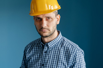 Portrait of caucasian young man engineer wearing yellow protecting safety helmet and shirt in front of blue wall isolated front view looking to the camera