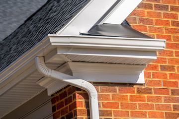 Closeup view of white gutter system with soffit vent, gutter guard, drop outlet, downspout, vinyl elbows, decorative trim molding, on the corner of a brick luxury house in America © tamas