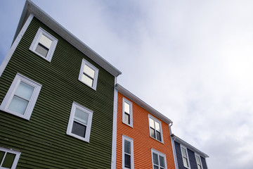 Blue, orange and green wood buildings with windows reflecting the cloudy sky. The roofs are flat with white trim. The sky is grey with some blue spots. 