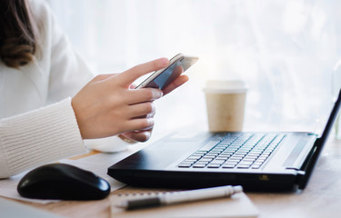 Close up of hand young female using mobile phone to searching information while she working with laptop on desk