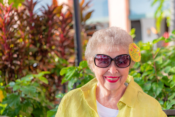 A senior woman at a restaurant in Hawaii, with a cute tropical drink umbrella in her hair. She wears sunglasses. - obrazy, fototapety, plakaty