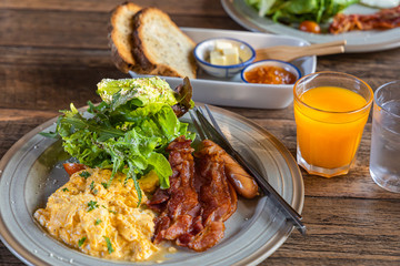 English breakfast-Scrambled eggs, bacon, vegetables, orange juice on wooden table.