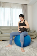 Pregnant young Vietnamese woman doing relaxing exercise on fitness ball at home