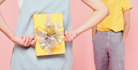 A woman in blue jean hiding a yellow present box behind her back and a woman in yellow blouse waiting for a gift, pink background