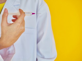 A woman doctor holding a syringe and needle of medication in right hand, standing on the yellow background (focus at the needle)