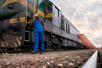 Freight train driver occupation. Caucasian bearded man standing by locomotive carrying cargo containers for shipping company. Railways transportation service.
