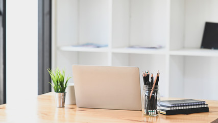 Computer laptop, Pencil holder, Potted plant, Notebook and coffee cup putting together on modern wooden table with office book shelf as background. © Prathankarnpap