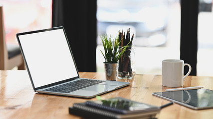 Laptop with white blank screen putting on modern wooden table together with computer tablet, coffee cup, potted plant, notebook. Orderly/Comfortable workplace concept.