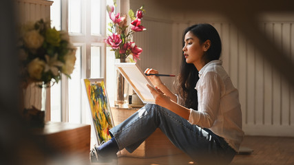 Photo of young artist girl holding a paint brush and drawing an oil colors on canvas while sitting...