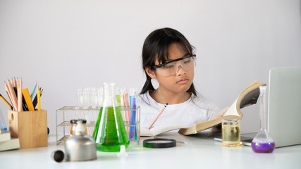 Photo of adorable schoolgirl reading a book while doing a scientific experiment at the white table with chemistry glassware over the white laboratory wall as background. Education for kids concept.