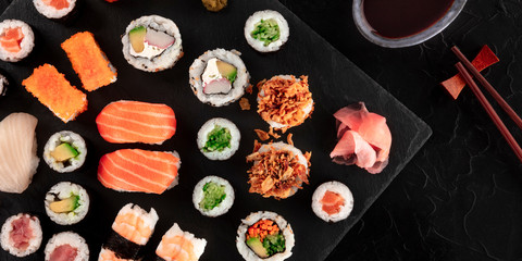 Large sushi set panorama with soy sauce and chopsticks, shot from above on a black background. An assortment of various maki, nigiri and rolls