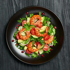 Shrimps salad with cherry tomatoes, cucumeber, avocado, lettuce and pomegranate on dish. Healthy seafood concept. Tasty grilled prawn shrimp and mix vegetable salad on black wood, top view.