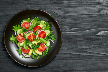 Fresh salad with cherry tomatoes, avocado,cucumeber and lettuce on dish. Healthy vegetarian meal concept. Tasty mixed leaves, tomatoes and mix vegetable salad on black wood, top view.