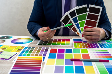 graphic or interior young designer in suit choosing color from swatch sample or catalogue palette guide at workplace. graphic designer with paint color palette samples at table