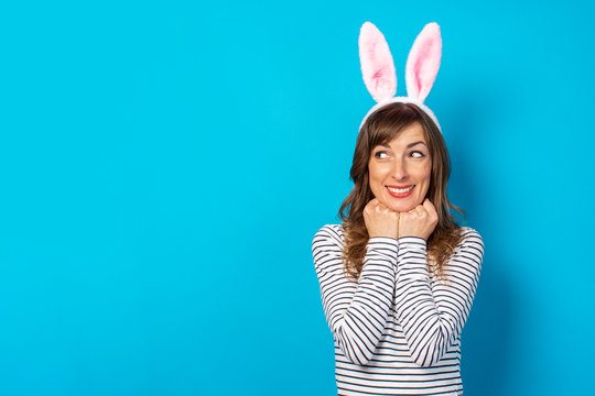 Joyful Young brunette woman with bunny ears and sincere feelings holds two hands under her chin on a blue background. Easter concept, surprise, hide and seek, emotional. Face expression. Banner