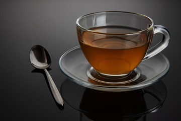 Glass cup of tea on a glass saucer with reflection, with a gradient of black and gray background....