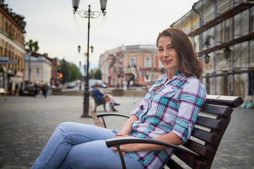 Fototapeta premium Brunette funny pretty girl sitting on bench in Central part of old city. Walk in downtown. Portrait of girl on street
