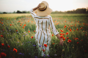 Young woman in rustic linen dress walking among poppy and cornflowers in summer meadow countryside...