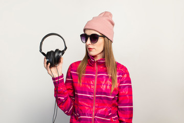 Friendly emotional. Young woman with glasses, a hat and a pink sports jacket holds headphones on a white background with copy space. Modern trendy music concept. Face expression. Banner