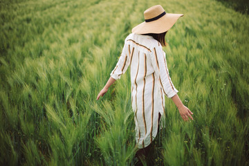 Young woman in linen dress and hat walking in green field of barley in countryside. Stylish girl in rustic dress enjoying peaceful moment in grass in summer. Tranquil rural moment. Copy space - Powered by Adobe