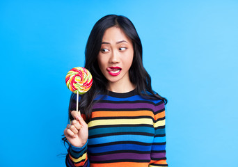 Young pretty woman lick her lips looking to big colorful lollipop against blue background.