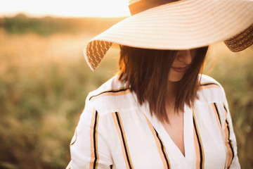 Portrait of beautiful woman in hat enjoying sunset golden light in summer meadow. Stylish rustic girl in linen dress relaxing in evening in countryside. Rural slow living. Atmospheric moment