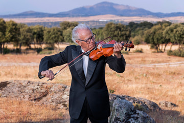 Elderly man, dressed in tuxedo, plays the violin in a natural environment