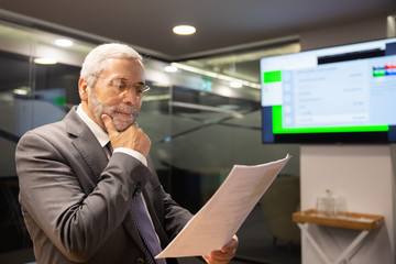 Focused mature businessman reading documents. Concentrated bearded worker in eyeglasses holding hand on chin and looking at annual report. Business concept