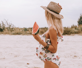 Young pregnant woman in a swimsuit and hat with a watermelon in hand on sand. pregnancy concept